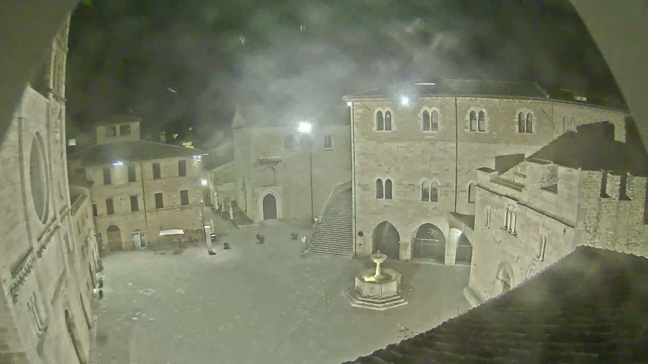 A nighttime view of a town square with several stone buildings surrounding a central fountain, under hazy conditions.