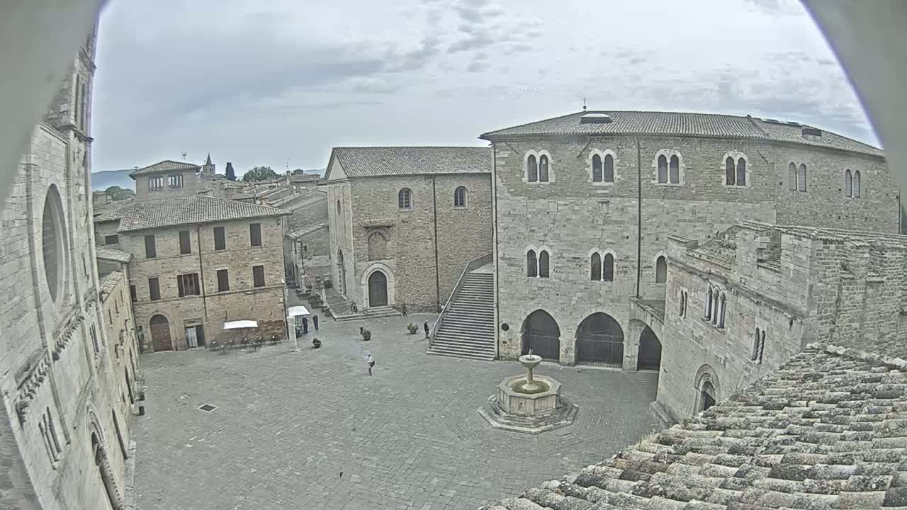 An overcast day reveals a stone plaza surrounded by medieval-style buildings, a fountain in the center, and a few people walking.