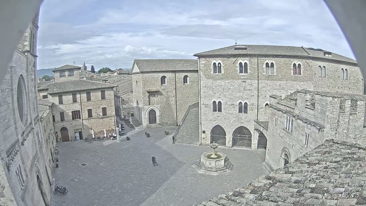 A stone plaza with a fountain is surrounded by medieval-style buildings under a partly cloudy sky.