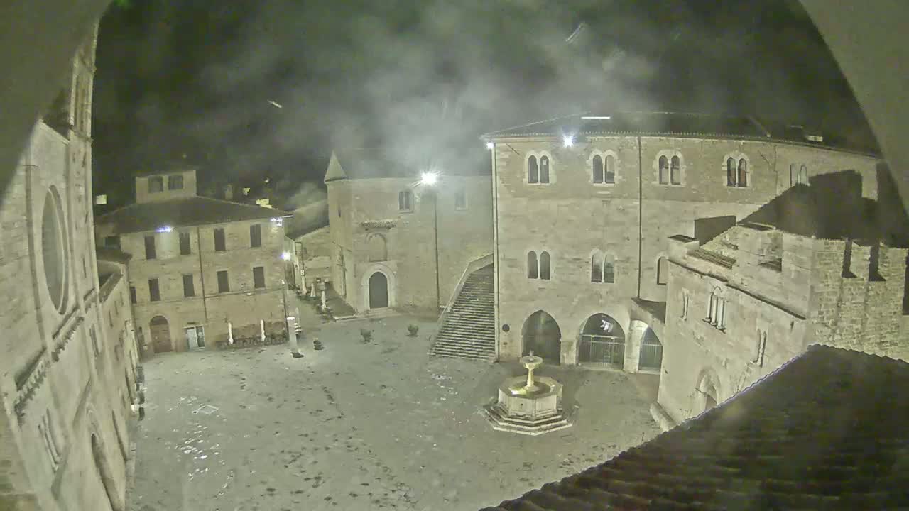 A nighttime view of a town square with stone buildings and a fountain, under a cloudy sky.