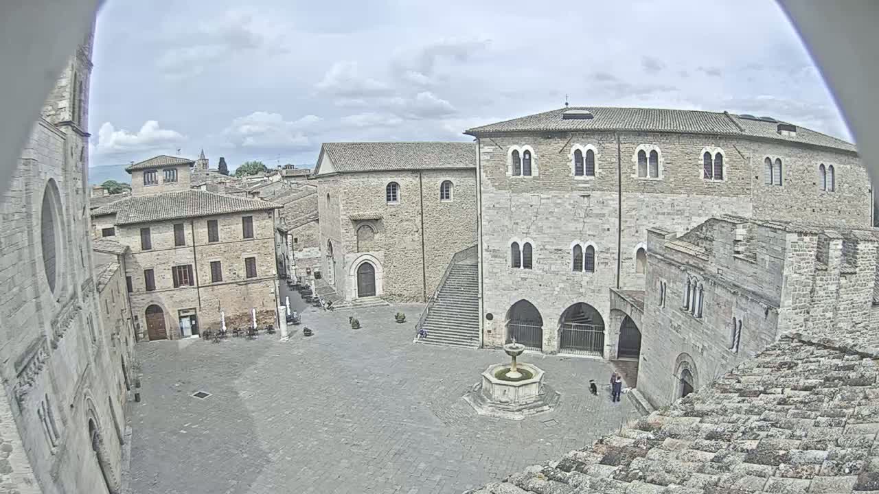 An overcast day reveals a stone plaza surrounded by medieval-style buildings, a fountain at its center, and a few people strolling.