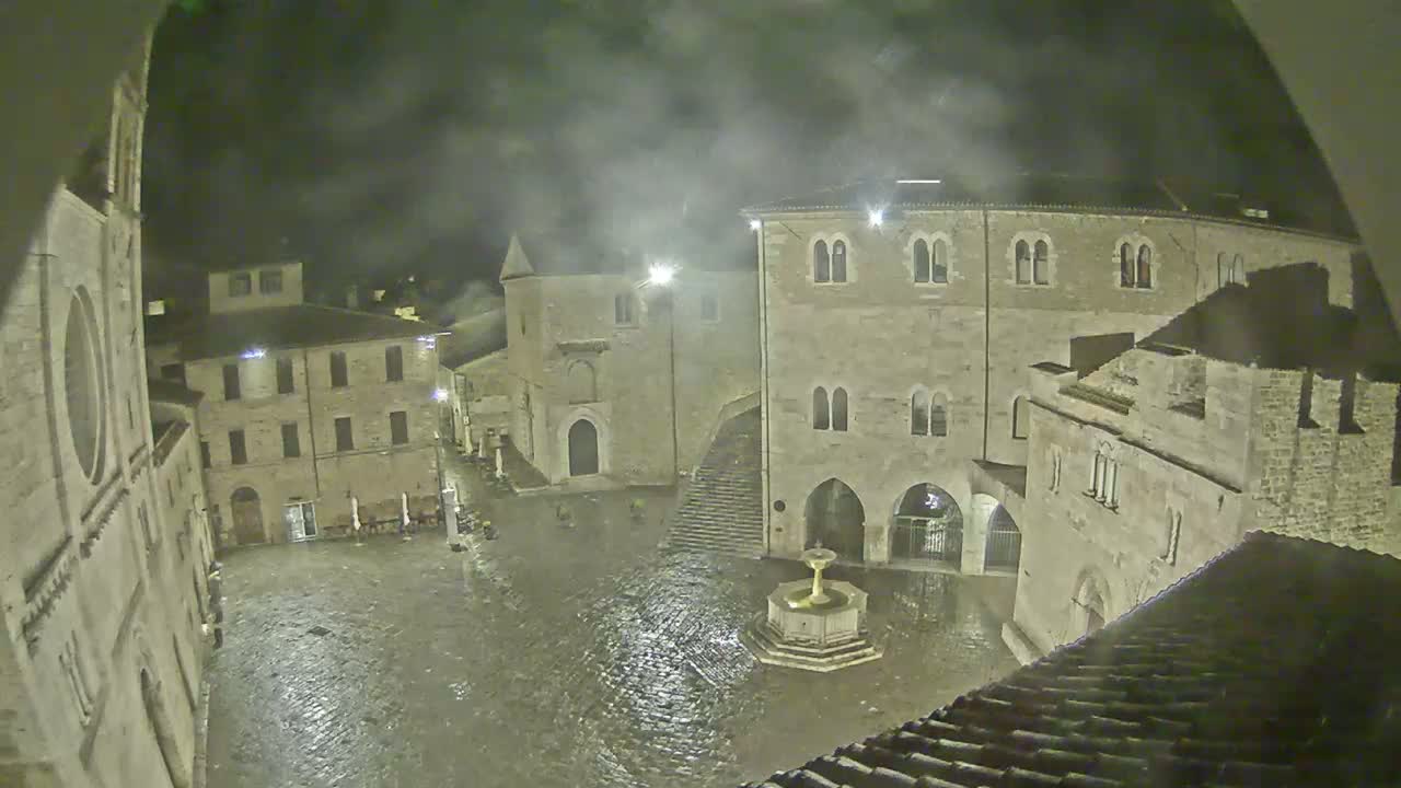 A rain-soaked town square at night, surrounded by ancient stone buildings, features a fountain in the center.
