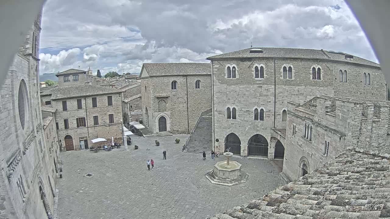 A high-angle view of a stone plaza in a historic town, under a cloudy sky, features a fountain in the center, several buildings, and a few people.