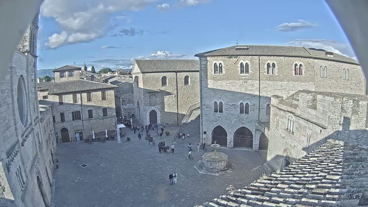 A high-angle view of a town square with stone buildings, a fountain, and a few people, under a partly cloudy sky.