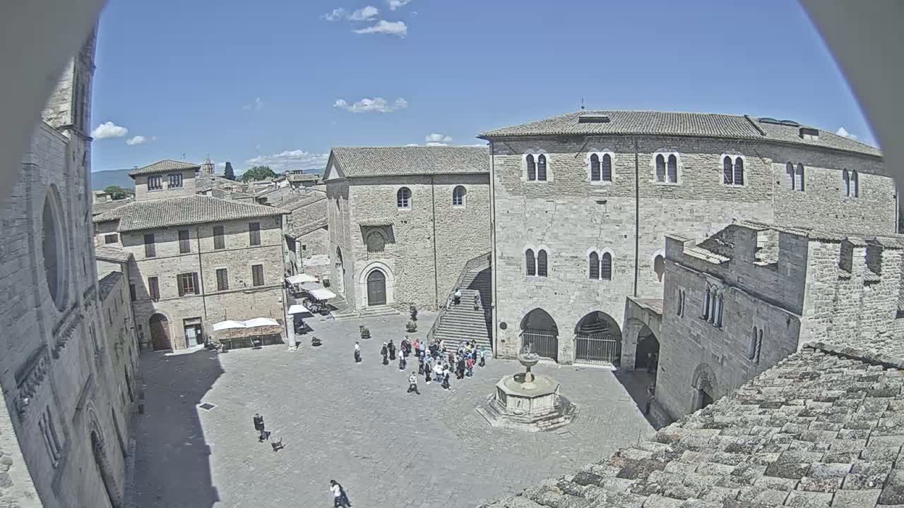 A sunny day reveals a town square in a historical European town, surrounded by stone buildings and featuring a fountain and a few people gathered in the center.