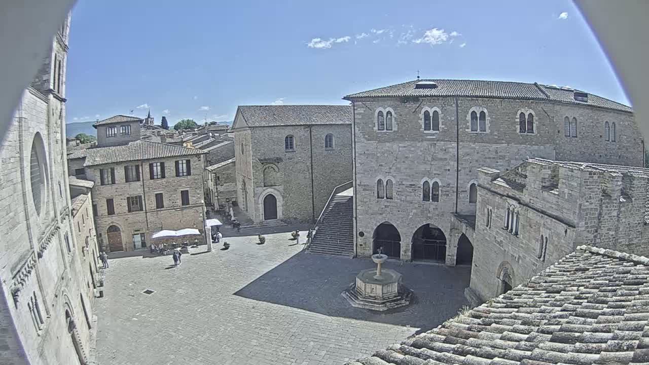 A sunny day reveals a cobblestone town square surrounded by medieval stone buildings, one featuring a prominent staircase, and a central fountain.