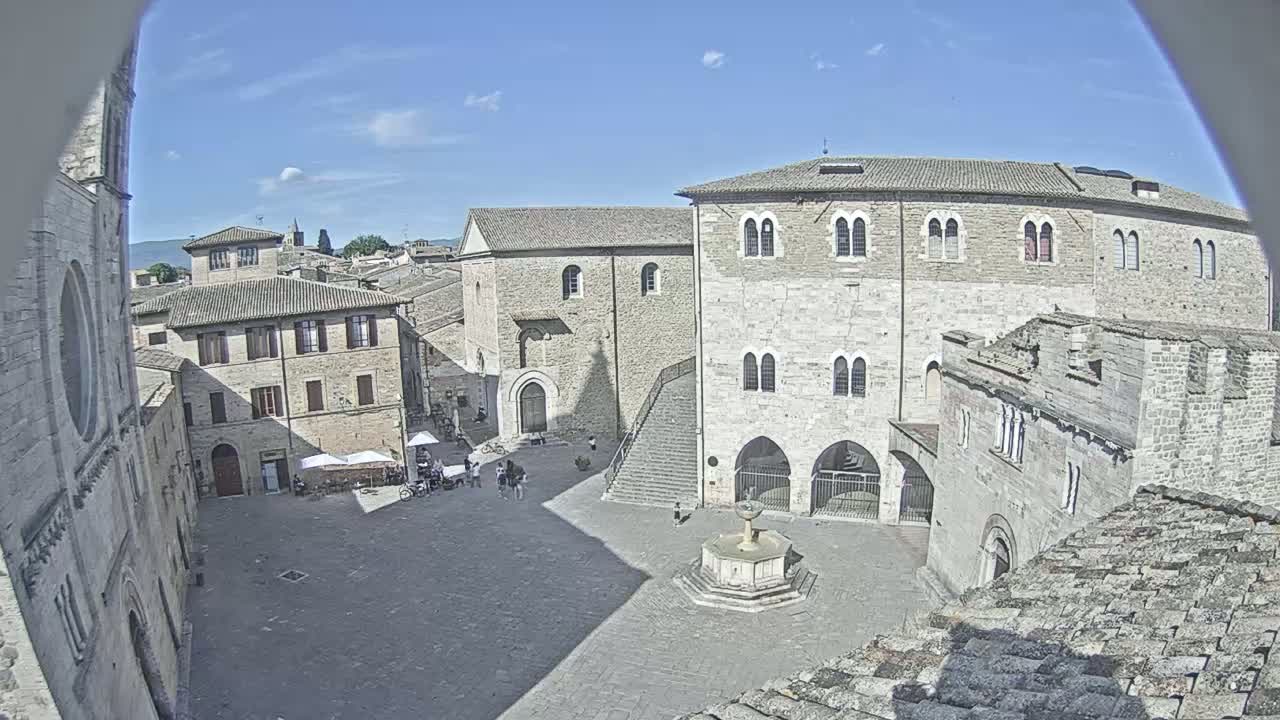 A sunny day reveals an open square surrounded by medieval stone buildings, featuring a central fountain and a few people.