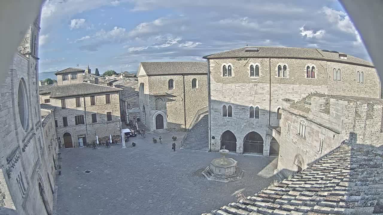 A high-angle view shows a town square with stone buildings, a fountain, and a few people, under a partly cloudy sky.