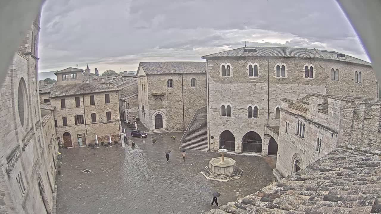 A wet, stone town square with several old buildings surrounding it, a fountain in the center, and a few people walking under a cloudy sky.