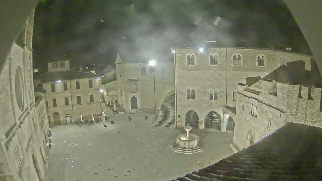 A nighttime view of a town square with several stone buildings surrounding a fountain, under hazy or foggy conditions.