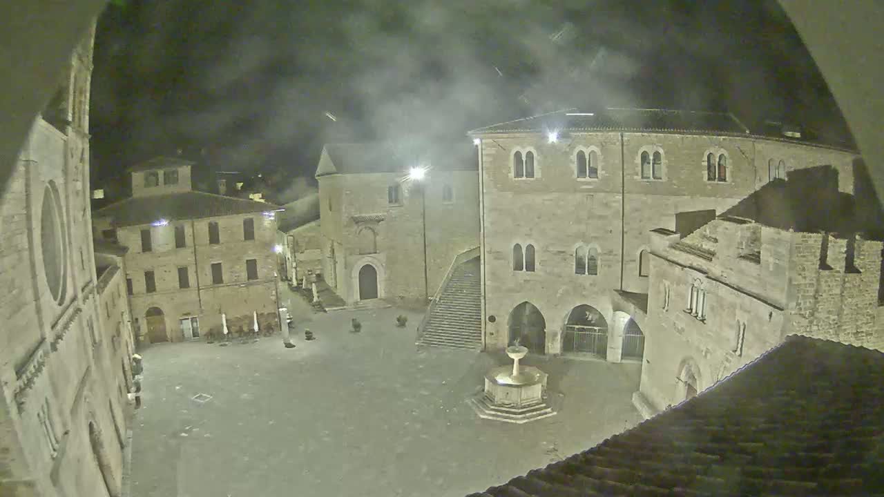 A nighttime view of a town square, surrounded by old stone buildings under a cloudy sky.