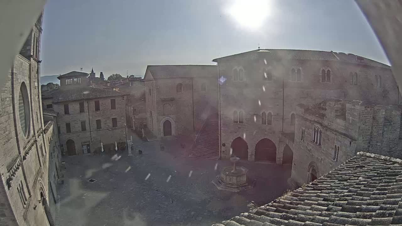 A sunny day reveals a town square surrounded by ancient stone buildings, a fountain, and a staircase leading up to a large structure.