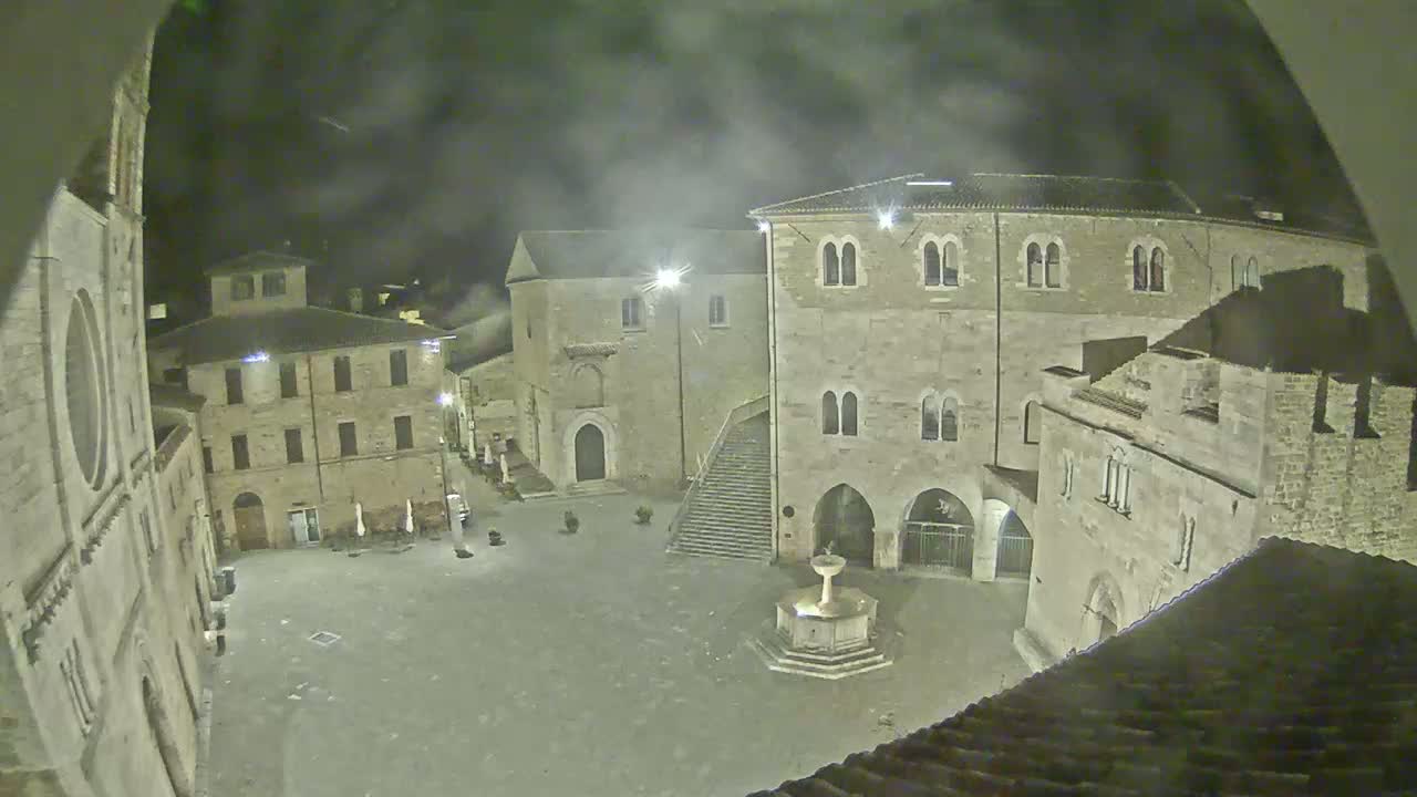 A nighttime view of a town square with stone buildings and a fountain, under hazy or foggy conditions.