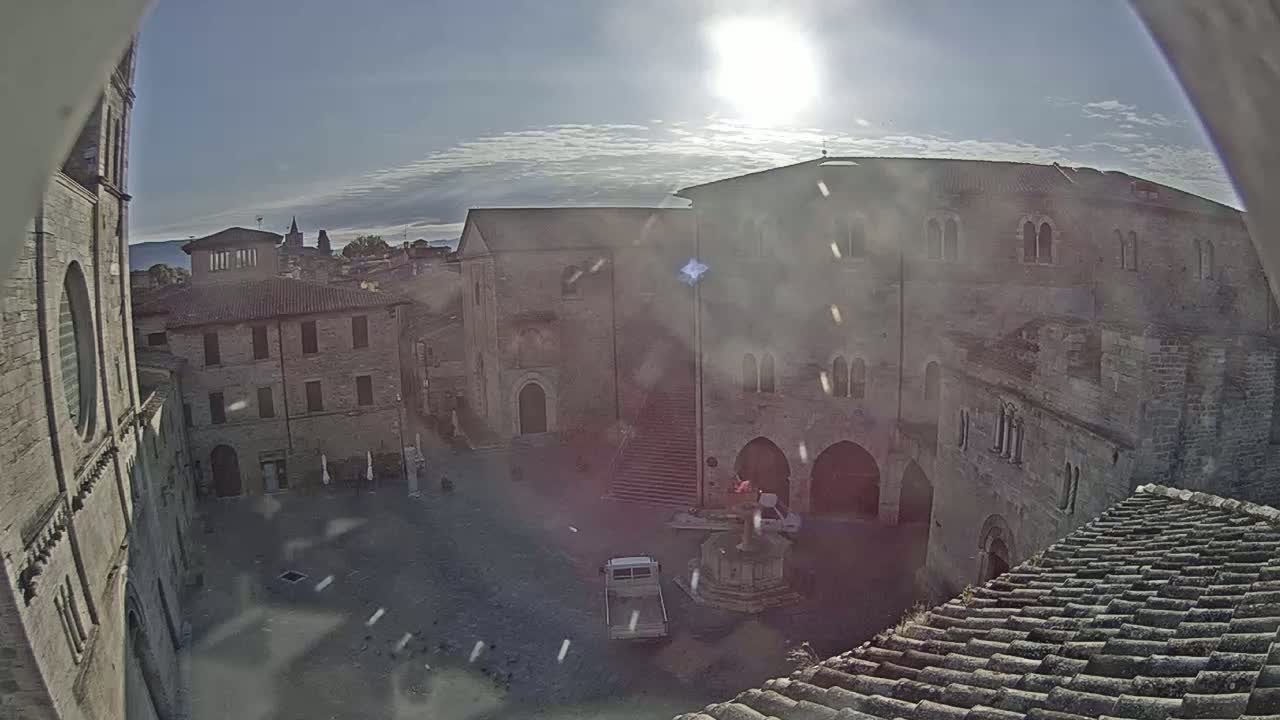 A sunny day reveals a high-angle view of a town square surrounded by aged stone buildings, one of which features a prominent staircase, and a small vehicle is parked nearby.