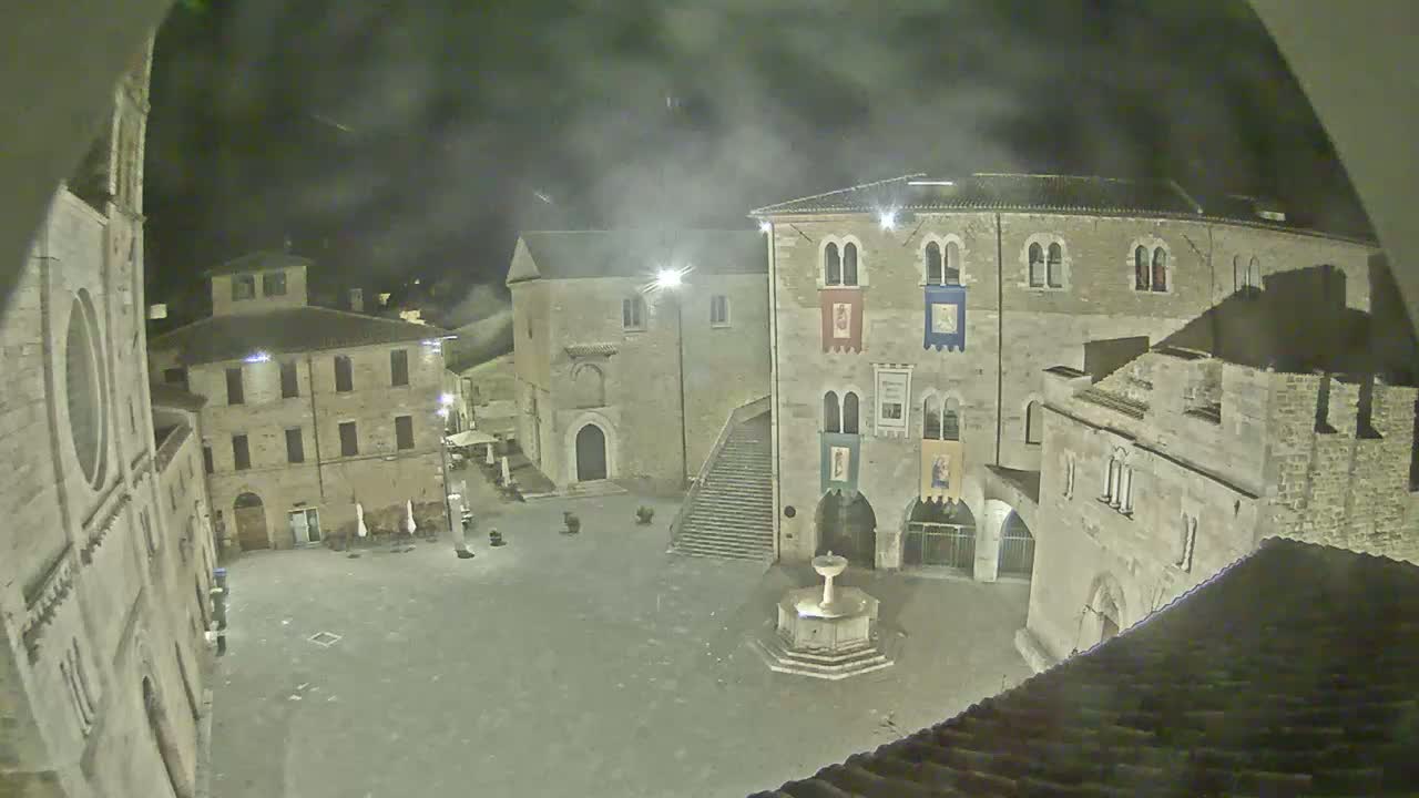 A nighttime view of a town square with stone buildings, a fountain, and a stairway, under a hazy sky.