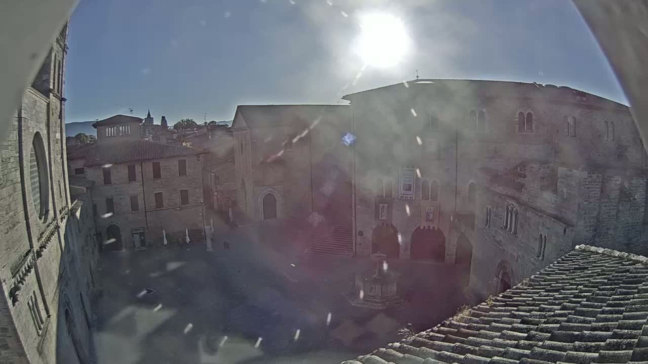 A sunny view overlooking a town square surrounded by aged stone buildings, one of which features a prominent staircase.