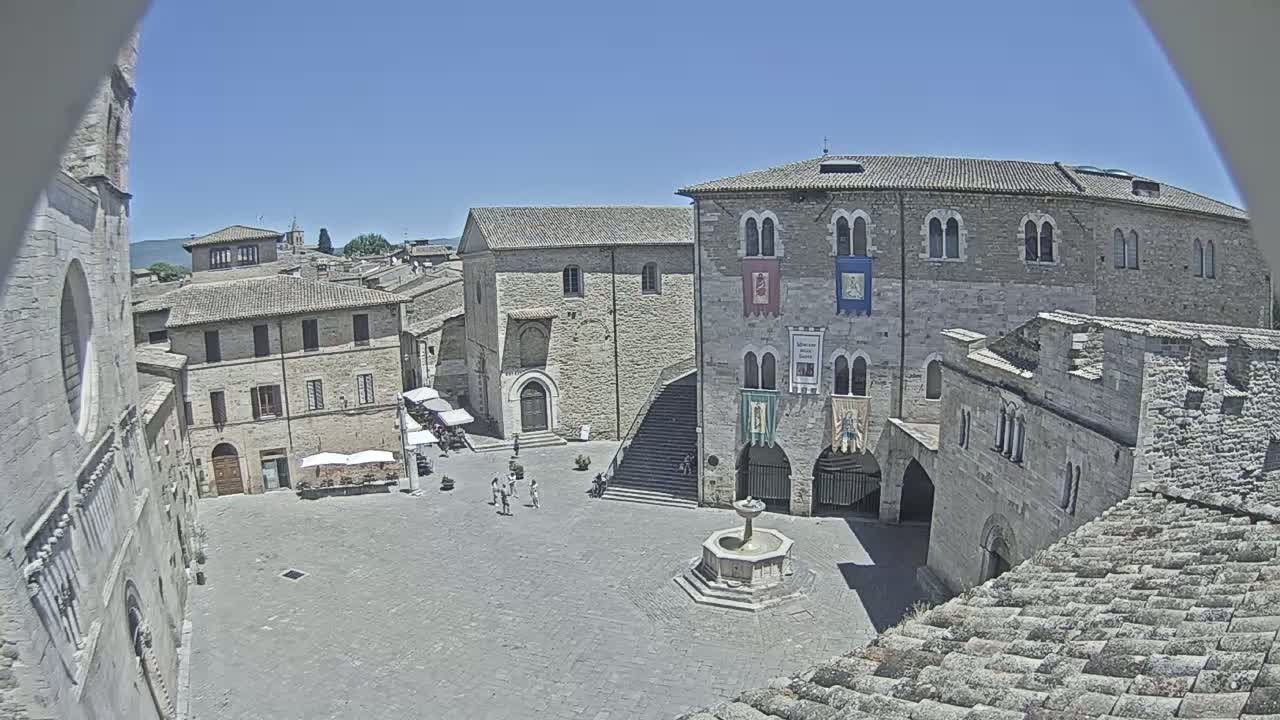A large, stone town square, under a clear blue sky, is surrounded by medieval buildings, one of which has a fountain in its center.