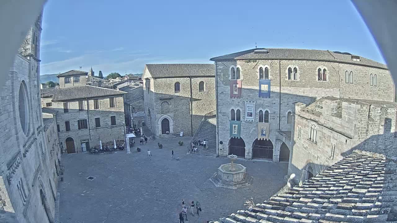 A sunlit town square in Italy, surrounded by medieval buildings, contains a fountain and a few people.