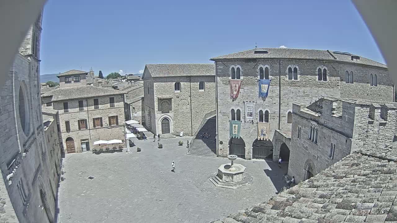 A town square with stone buildings, a fountain, and a few people is viewed from above on a sunny day.