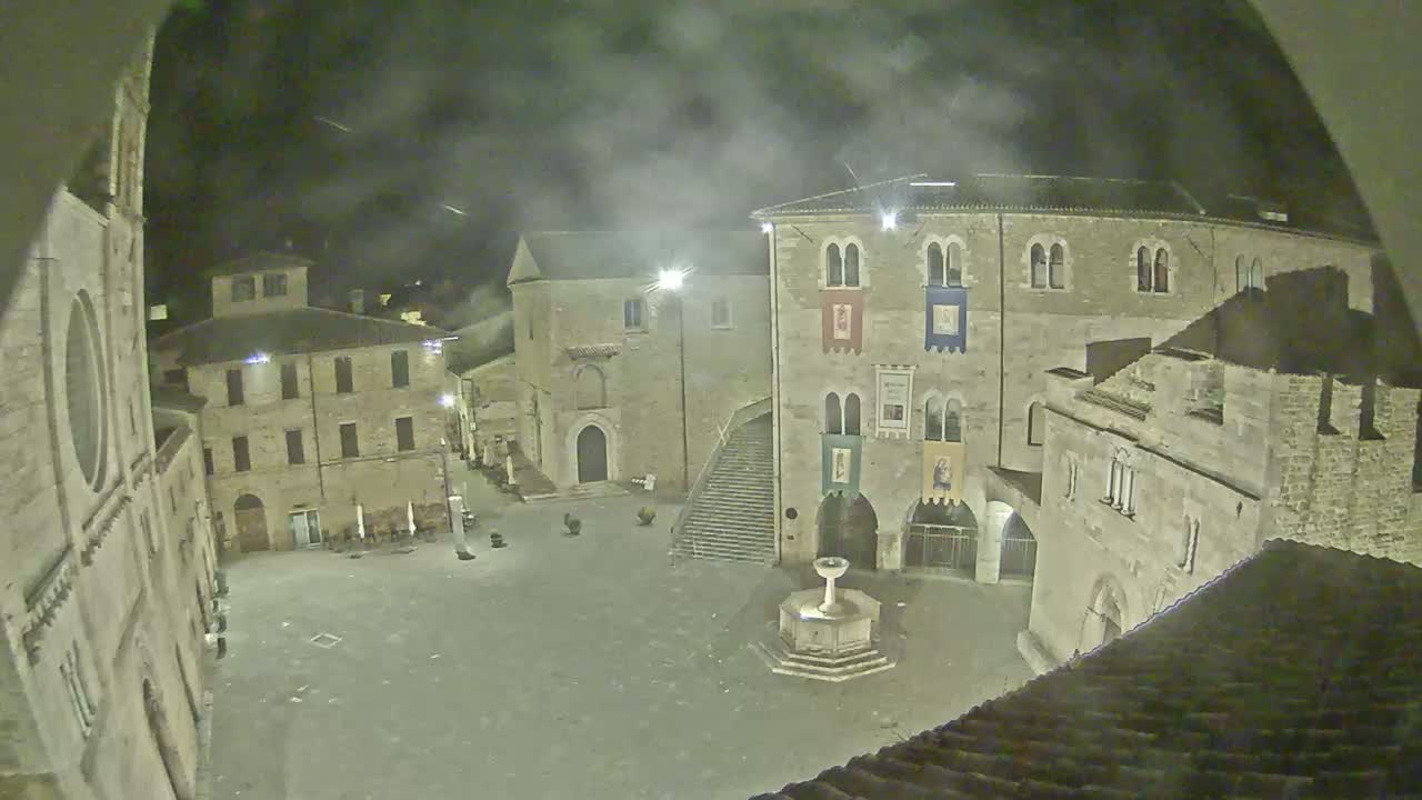 A nighttime view of a stone plaza with several buildings, including a church and a building with banners, under a cloudy sky.