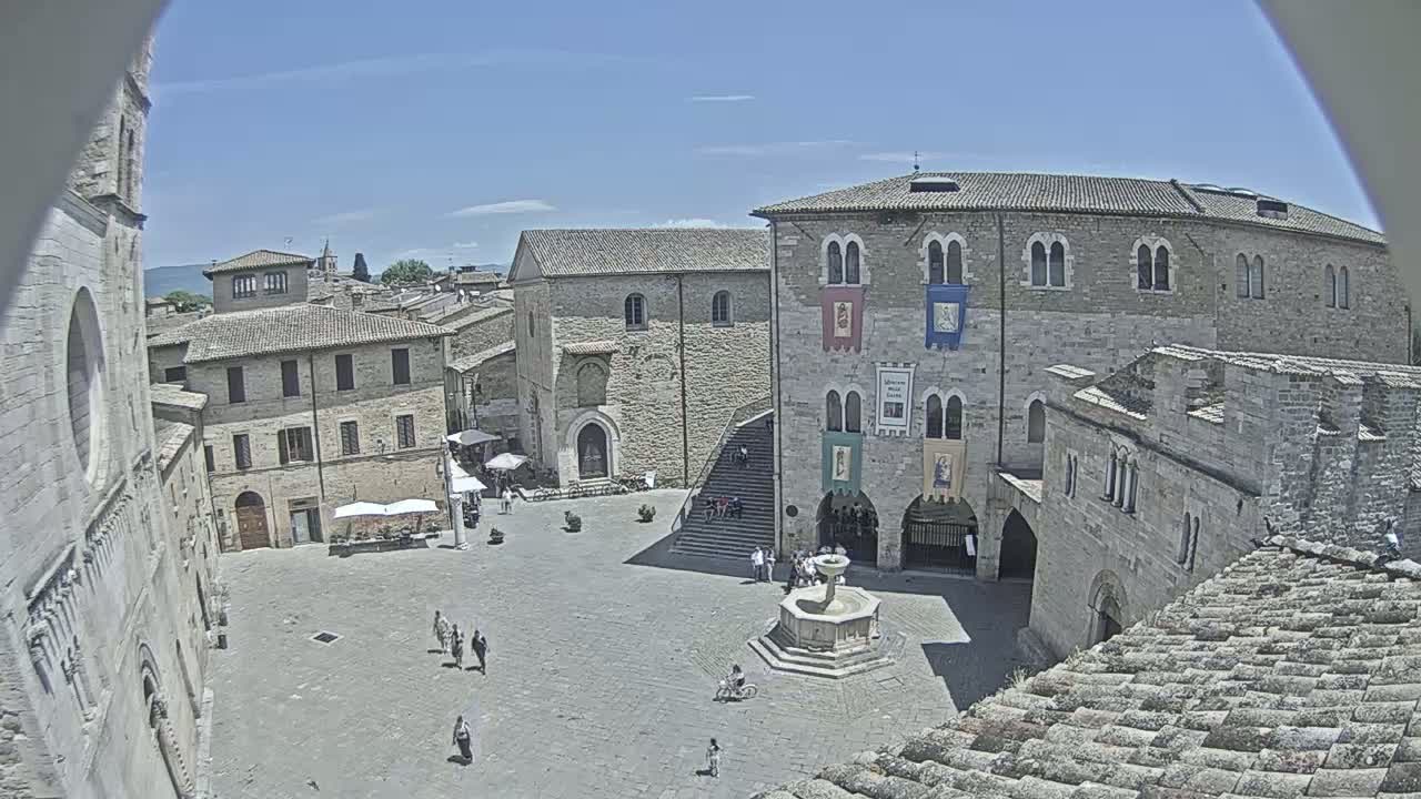 A high-angle view shows a town square with stone buildings, a fountain, and a few people under a sunny sky.