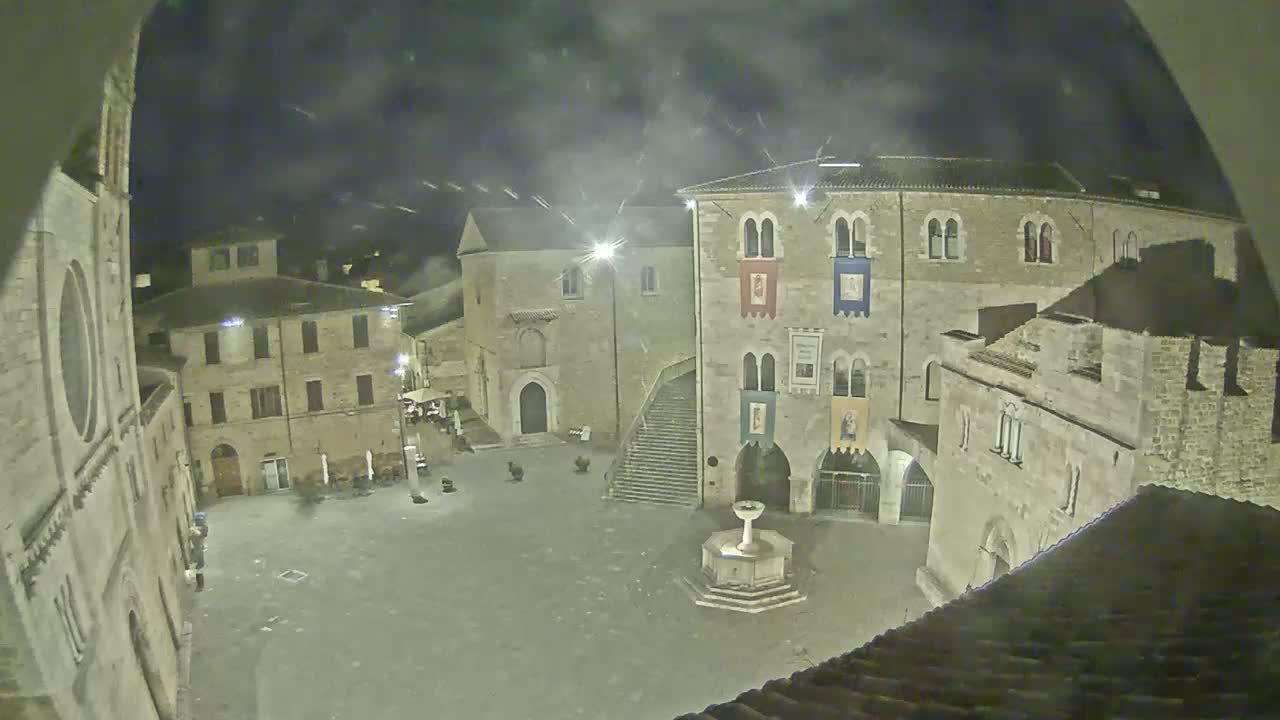A nighttime view of a town square with stone buildings, a fountain, and a partly cloudy sky.