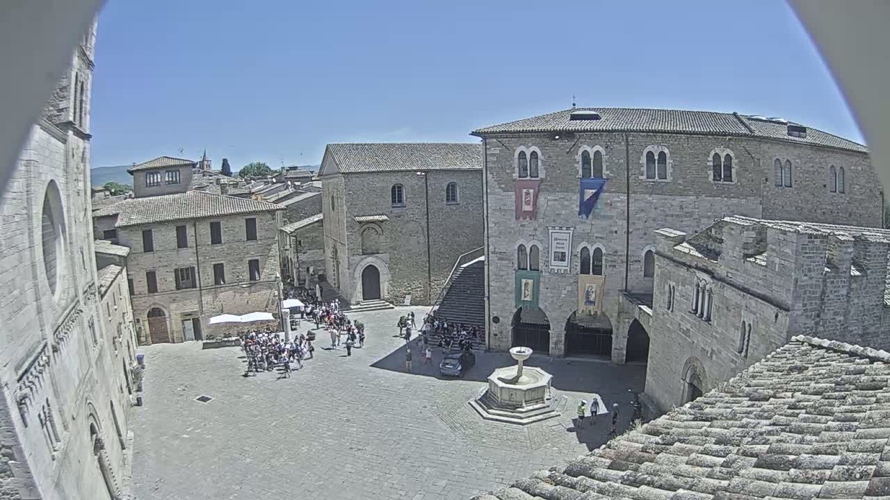 A sunny day reveals a large town square in a historic European city, where a crowd gathers around a fountain situated between ancient stone buildings.