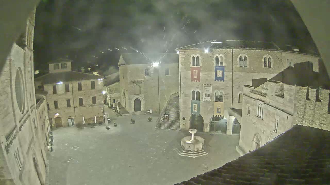 A nighttime view of a town square, under a slightly hazy sky, with stone buildings surrounding a central fountain.