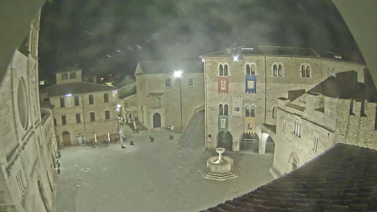 A nighttime view of a town square with stone buildings, a fountain, and a partly cloudy sky.