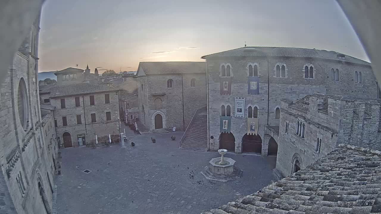 A stone plaza with a fountain, surrounded by medieval-style buildings, is seen at sunrise under a mostly clear sky.
