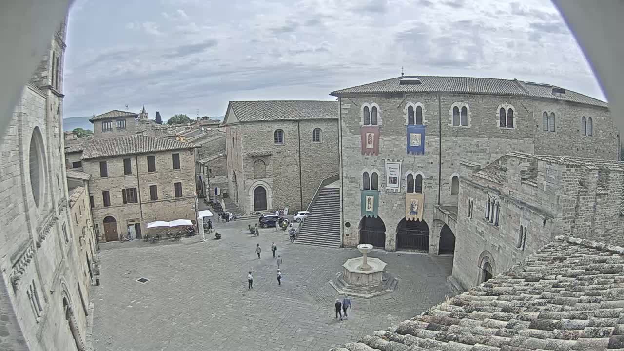 A town square with stone buildings, a fountain, and a few people, under a partly cloudy sky.