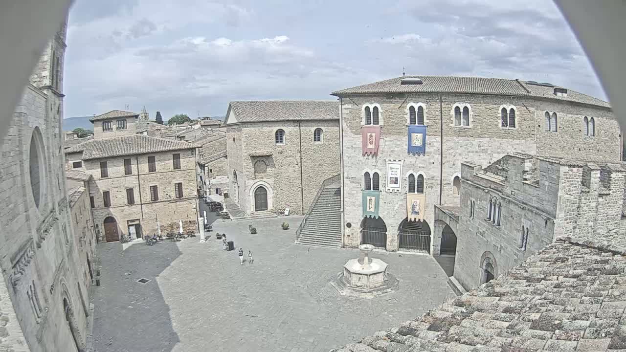 A mostly sunny day reveals a town square surrounded by stone buildings, one featuring a staircase and a fountain at its center.