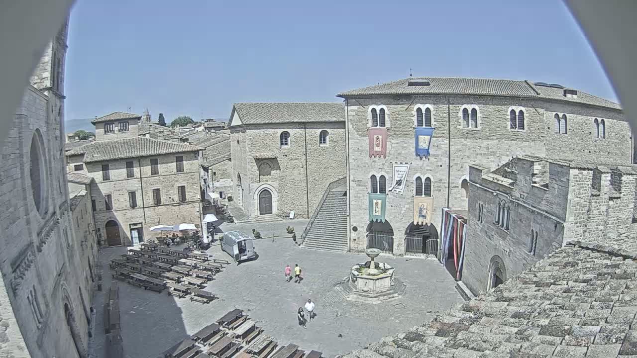 A sunny, high-angle view of a large town square in a medieval-style town, featuring stone buildings, a fountain, and several people.