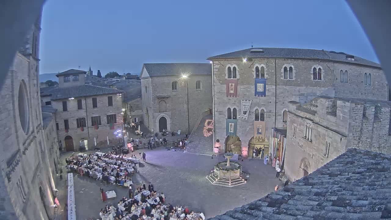 A town square at twilight is filled with people seated at long tables for a meal, surrounded by medieval-style buildings, one of which has banners hanging from its facade.