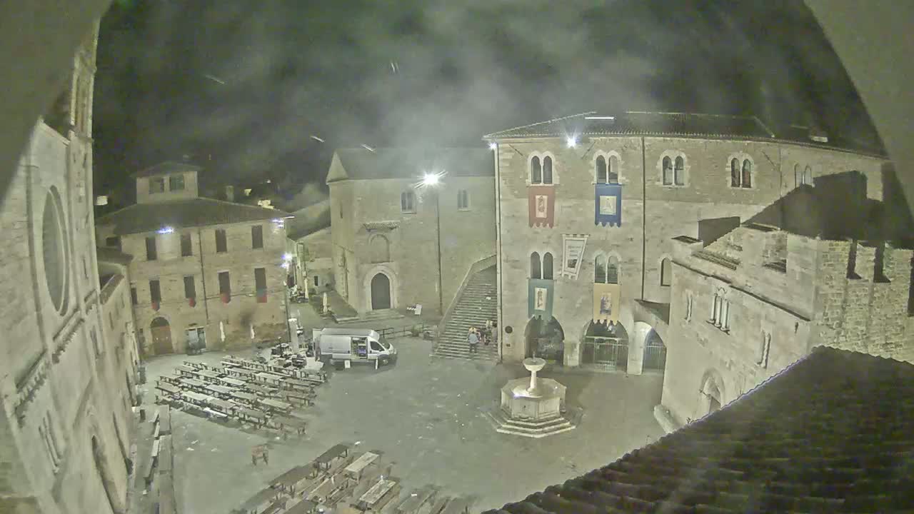 A nighttime view of a town square with stone buildings, a fountain, and several tables set up, under a hazy sky.