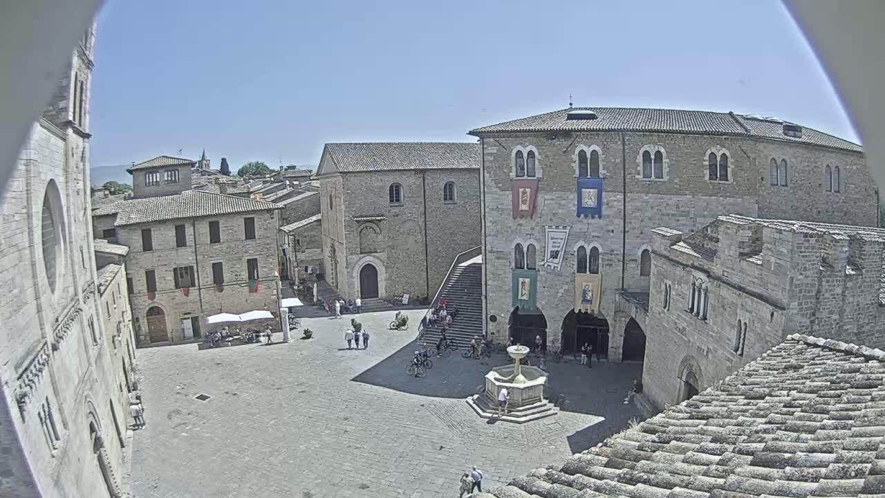 A high-angle, sunny view of a town square in a historic European village, centered around a fountain with several people and bicycles, surrounded by stone buildings.