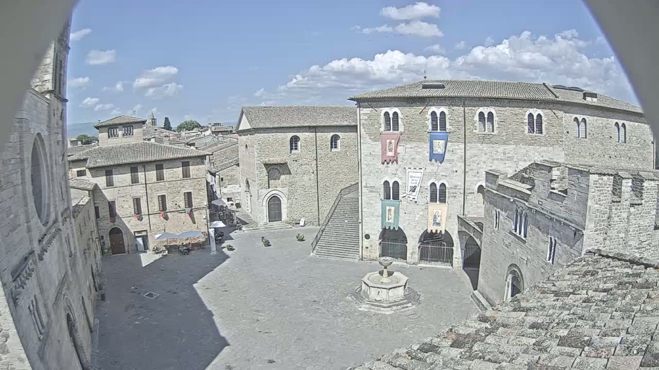 A sunny day reveals a European town square with stone buildings, a fountain, and a stairway leading to an upper level.