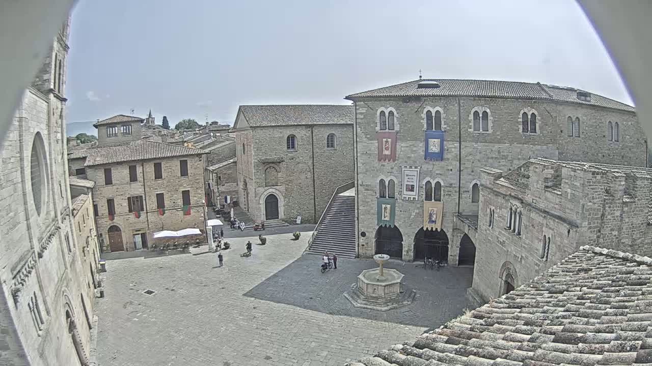 A high-angle view of a town square on a sunny day, centered around a fountain and surrounded by medieval-style stone buildings.