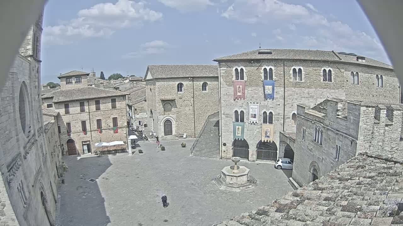 A mostly empty town square with stone buildings, a fountain, and a staircase under a partly cloudy sky.