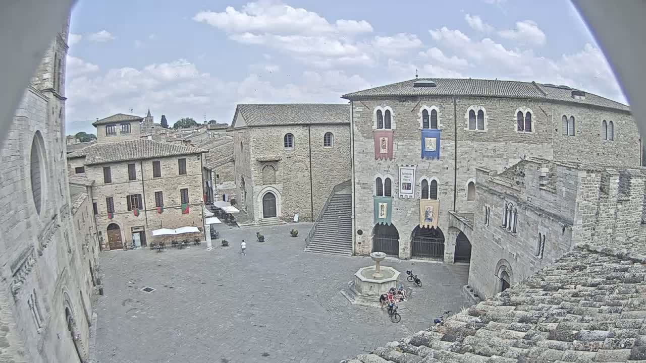 A mostly sunny day overlooks a town square with several stone buildings, a fountain, and a few people and bicycles.