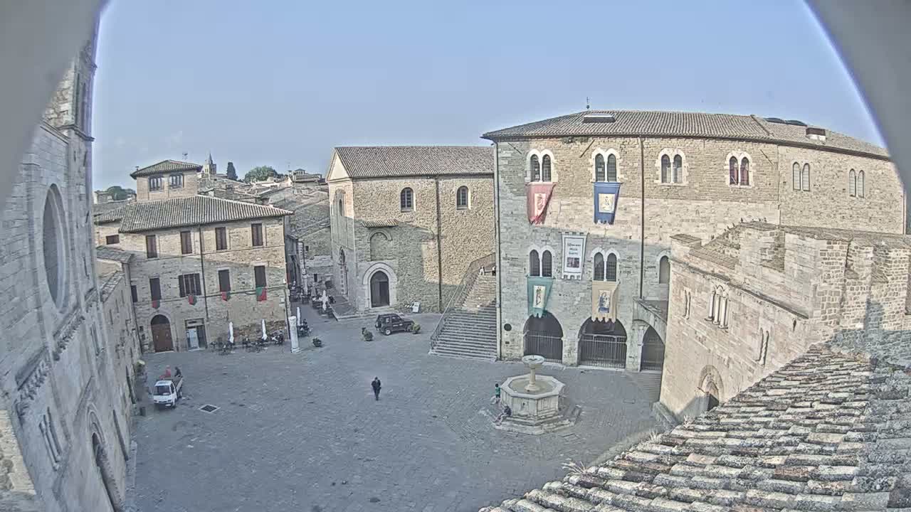 A high-angle view shows a town square surrounded by stone buildings under a clear, sunny sky, with a few people and vehicles visible.