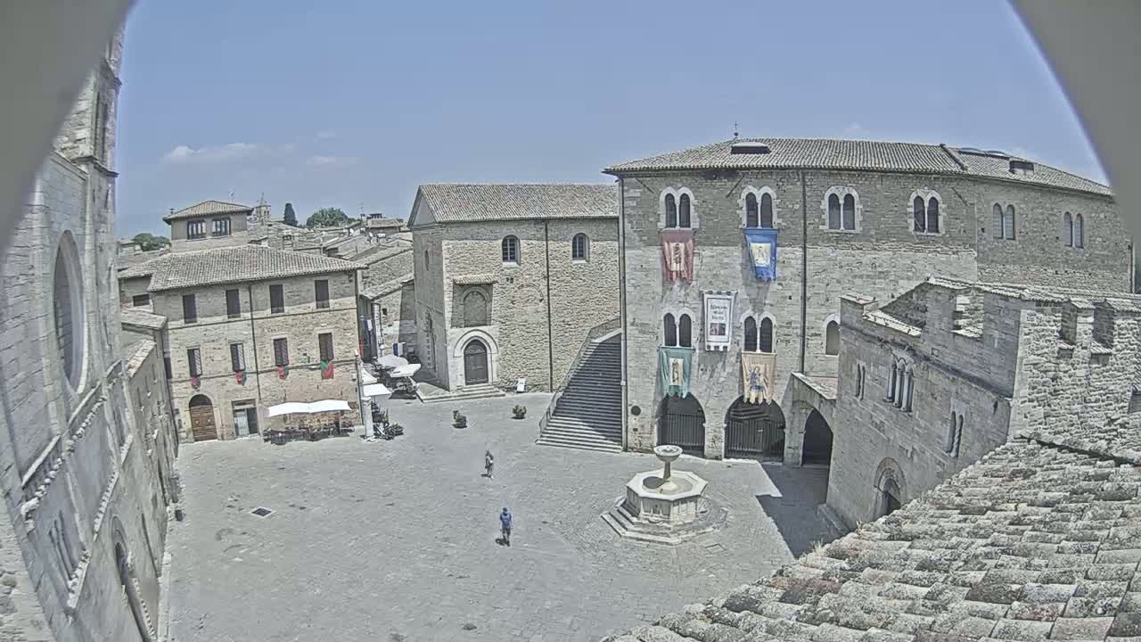A mostly empty town square with a stone fountain, surrounded by medieval-style buildings under a clear blue sky.
