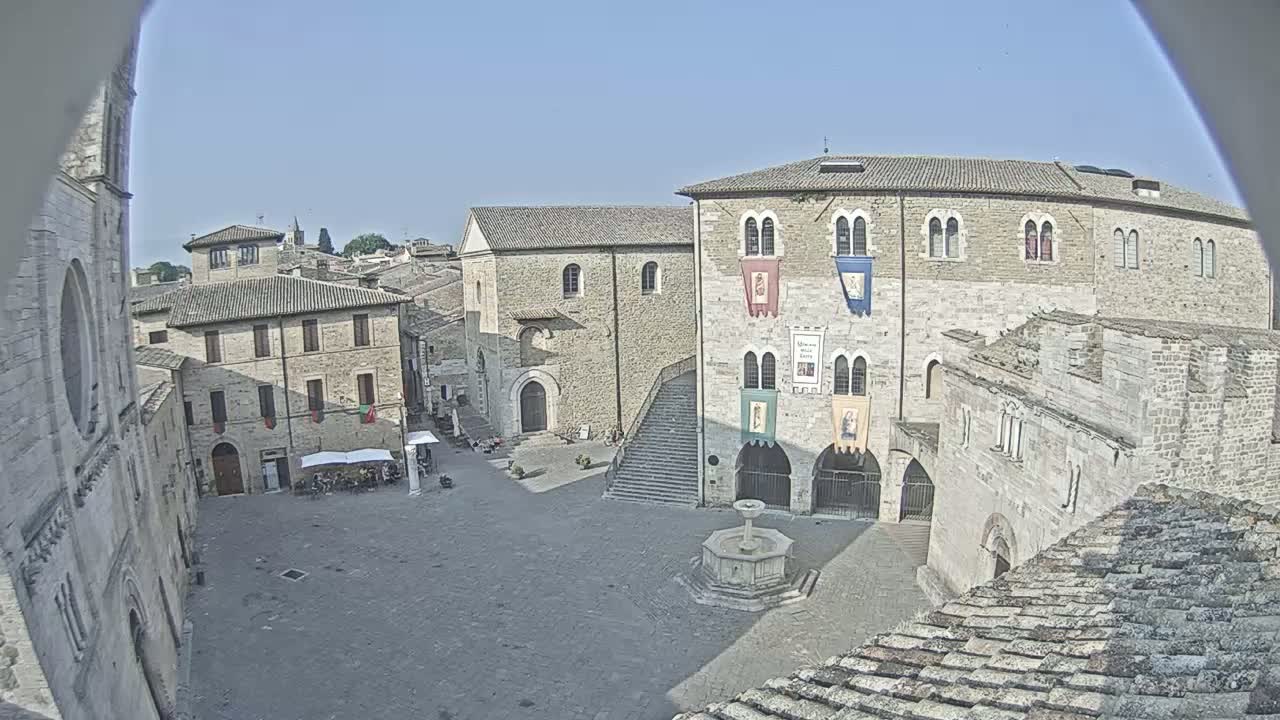 A large, stone town square with a fountain in the center, surrounded by medieval-style buildings under a clear, sunny sky.
