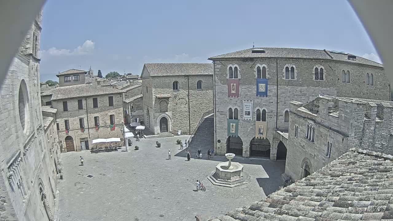 A sunny day overlooks a town square with several stone buildings surrounding a fountain, some people are present.