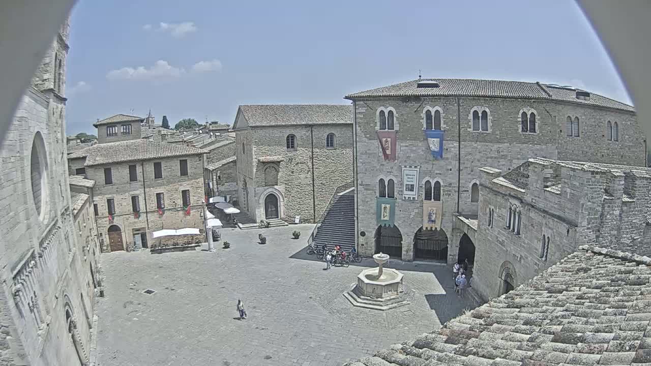 A town square with stone buildings, a fountain, and a few people, under a clear blue sky.