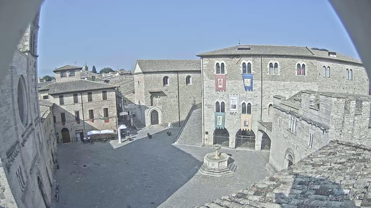 A high-angle, sunny-day view of a town square in a historic European town, featuring several stone buildings, a central fountain, and a staircase leading to a large building with banners.