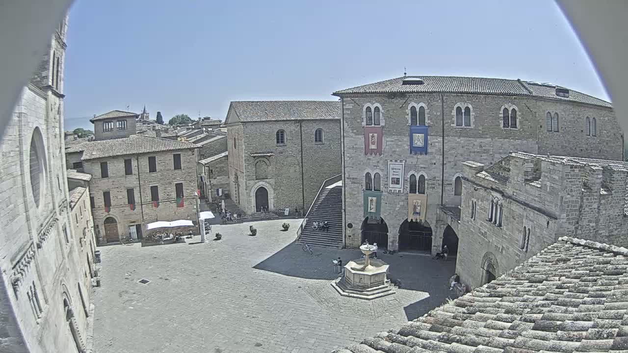 A sunny view of a town square in a European-style town, featuring stone buildings, a fountain, and a staircase leading up to a large building.