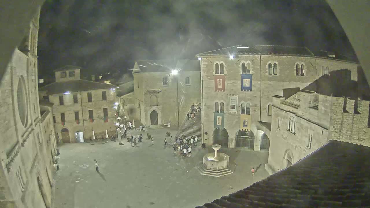 A high-angle view of a town square on a sunny day, featuring several stone buildings surrounding a fountain and a few people scattered throughout the space.