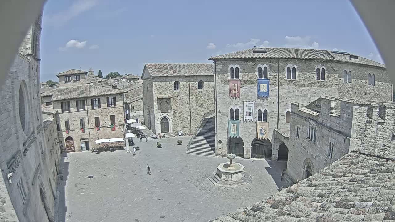 A mostly sunny day reveals a large, paved town square surrounded by ancient stone buildings, with a fountain at its center.