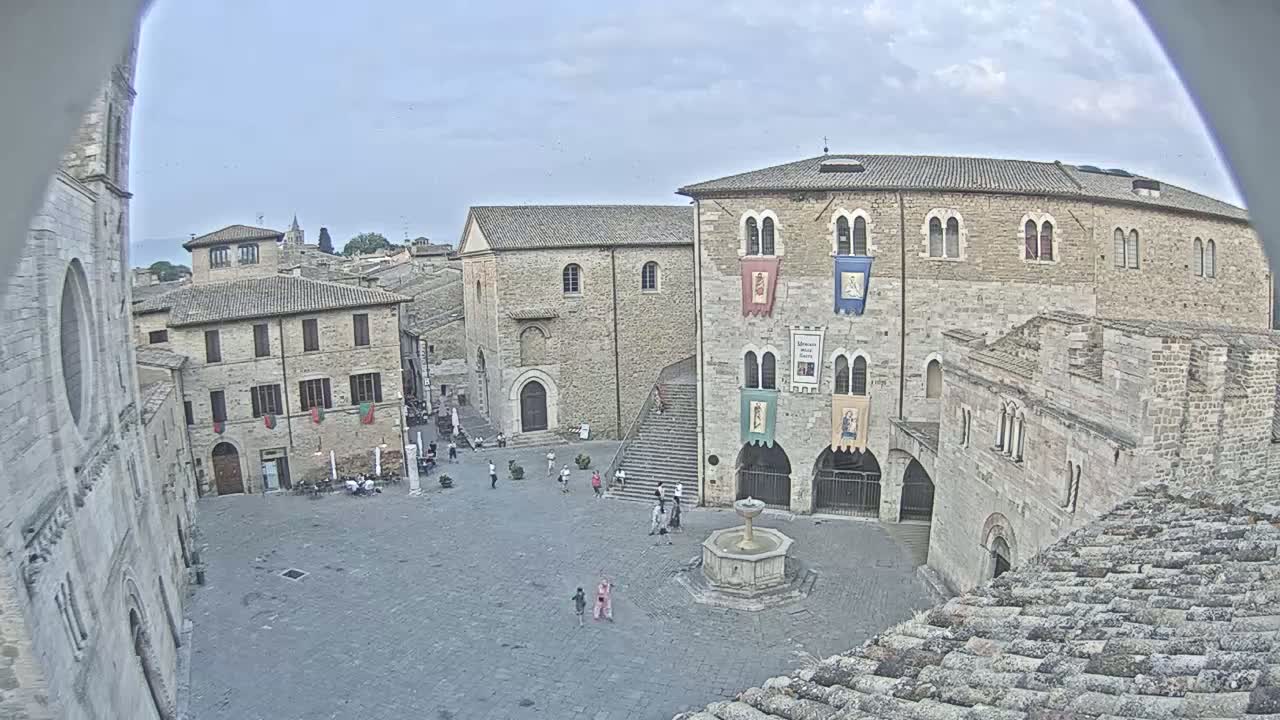 A mostly empty town square, surrounded by stone buildings under a partly cloudy sky, features a fountain in its center.